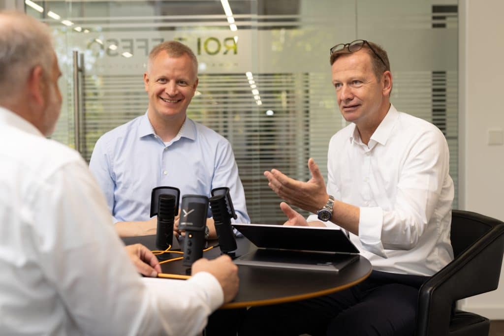Drei Geschäftsmänner sitzen an einem runden Tisch in einem modernen Büro mit Glaswänden. Sie führen ein professionelles Gespräch, während zwei Mikrofone auf dem Tisch aufgestellt sind, was auf eine Podcast- oder Aufnahmesession hindeutet. Einer der Männer spricht gestikulierend, während die anderen aufmerksam zuhören. Im Hintergrund spiegelt sich das Firmenlogo auf der Glaswand, was eine professionelle und kooperative Arbeitsumgebung vermittelt.